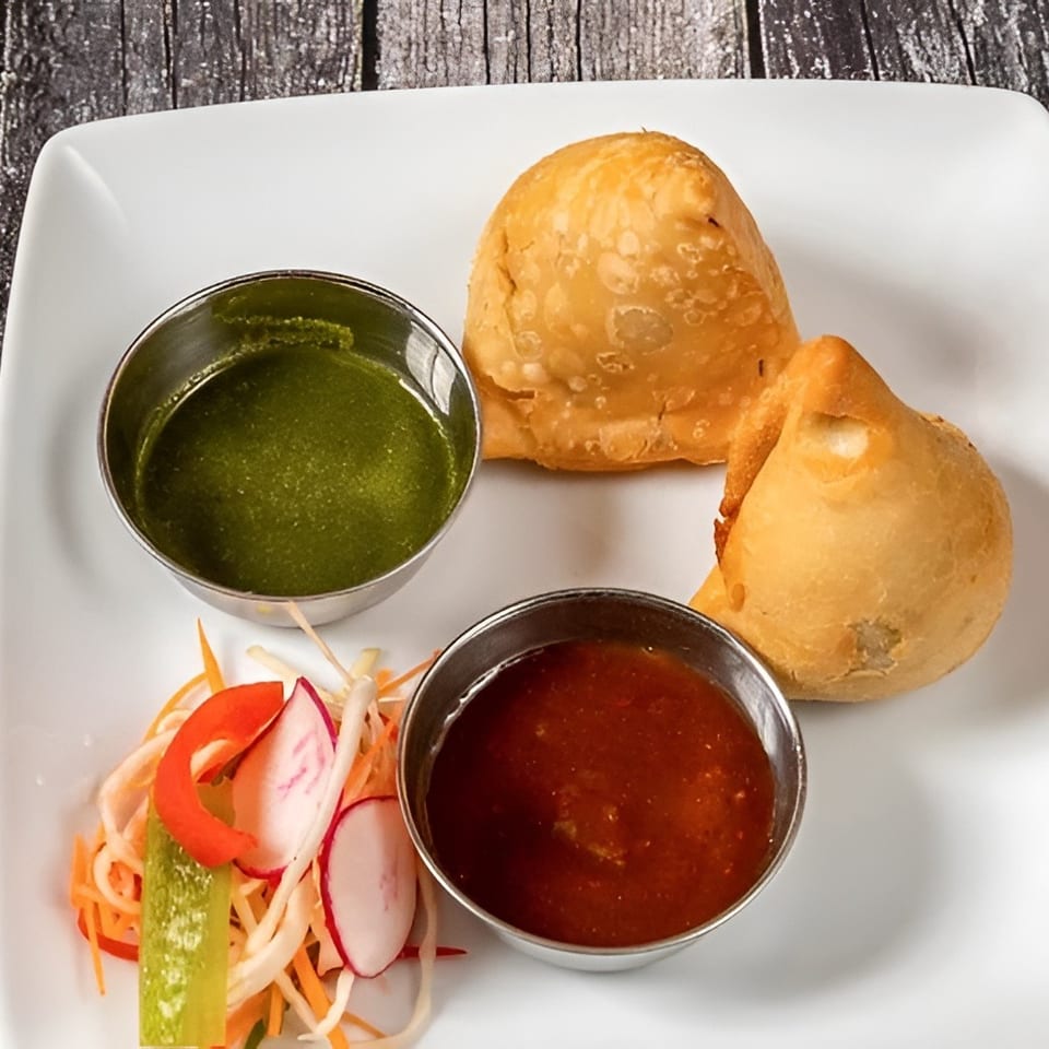 Two golden-brown samosas with green and red dipping sauces and a side salad on a white plate.