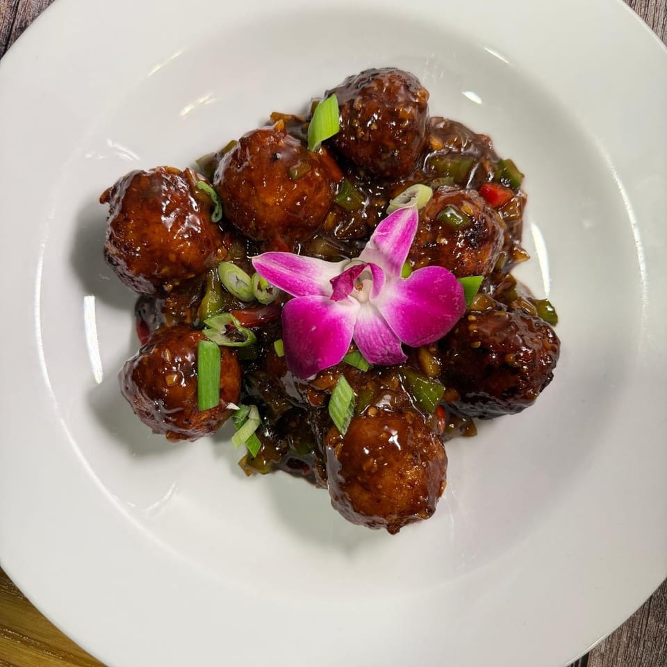 Brown meatballs in a dark sauce with green onions and a purple orchid on a white plate.