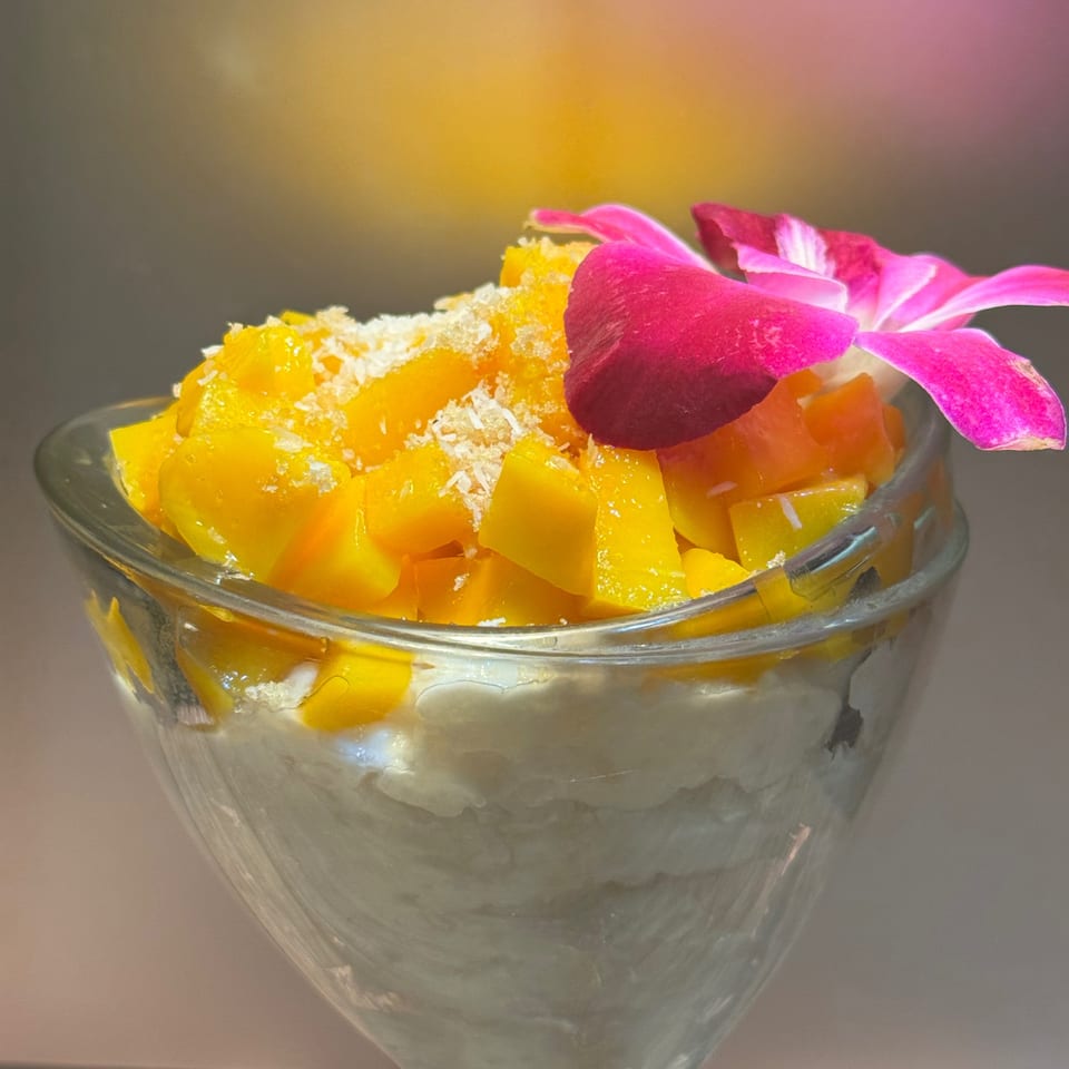 A glass of mango sticky rice dessert, topped with shredded coconut and a pink orchid.